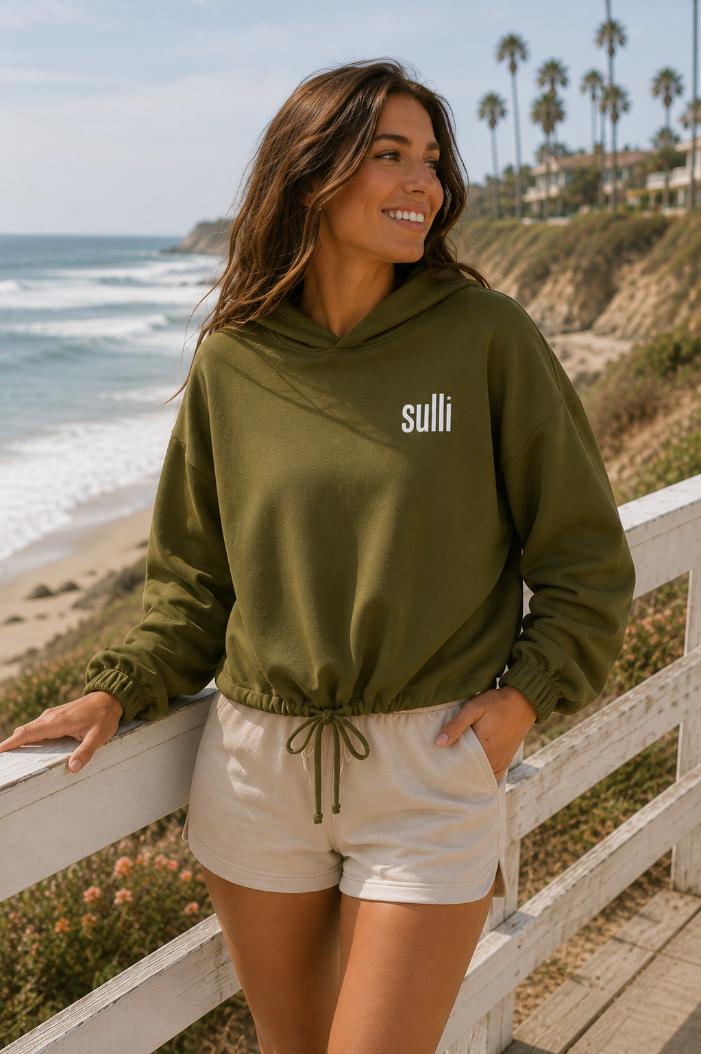 A woman wearing an olive green hoodie with 'suli' printed in white on the chest, paired with cream shorts, posing on a wooden boardwalk overlooking a beach and ocean.