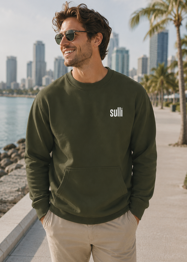 A man wearing a Sulli olive green crewneck sweatshirt with a white logo, standing along a sunny waterfront with palm trees and a modern city skyline.