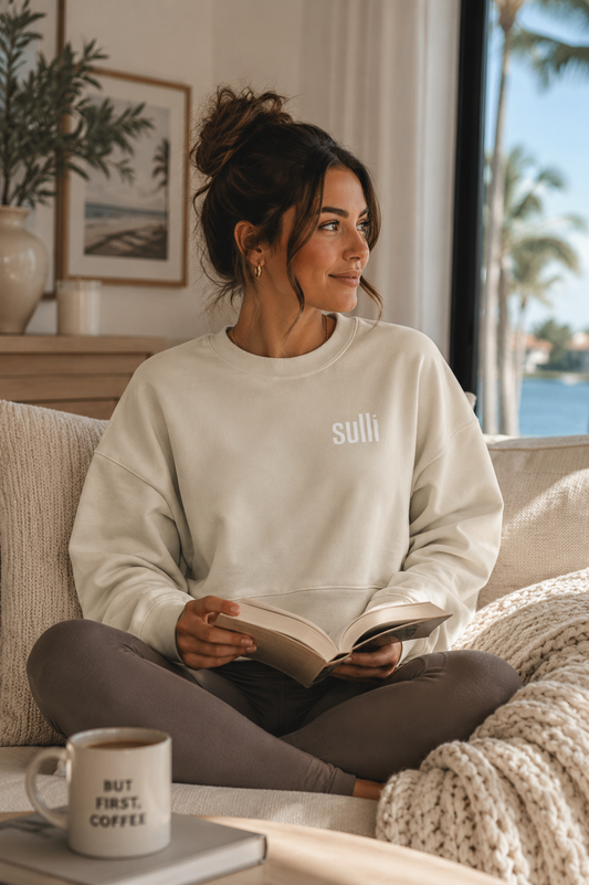 A woman sits cross-legged on a beige couch in a bright living room, wearing a cream sweatshirt with the brand name 'sulli' printed in white on the chest, paired with dark gray leggings. She is holding an open book, with a mug in the foreground and a window showing a sunny outdoor view in the background.