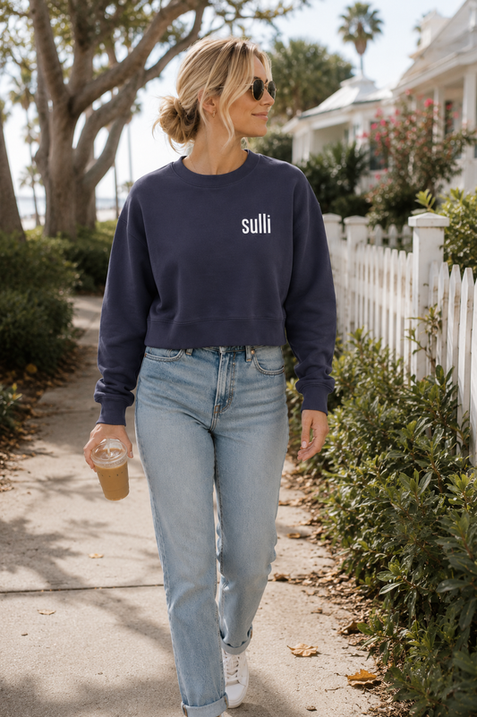 A woman walking outdoors on a sunny day wearing a navy blue crewneck sweatshirt with the word 'sulli' printed in white on the chest, paired with light-wash blue jeans and white sneakers.