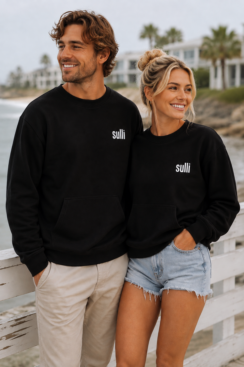 A man and a woman standing together on a beach boardwalk wearing matching black crewneck sweatshirts with the white text 'sulli' on the left chest. The man wears beige pants, and the woman wears light-wash denim shorts.