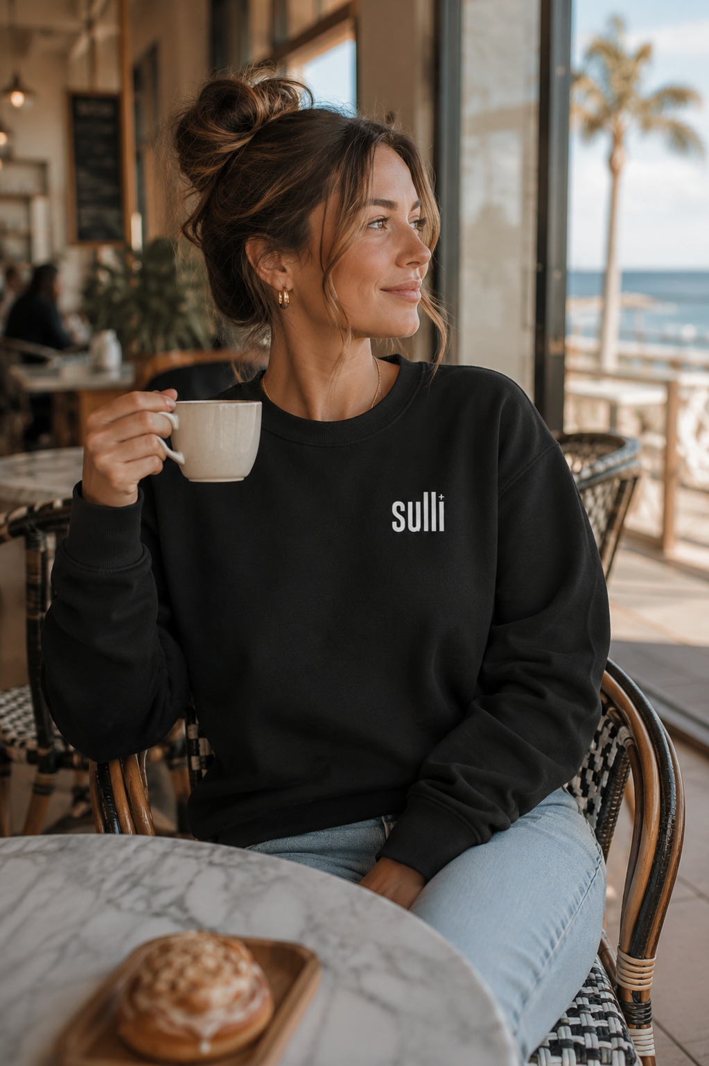 A woman sitting in a cafe chair by a window, holding a white mug. She is wearing a black crewneck sweatshirt with the word 'sulli' printed in white on the left chest, paired with light blue jeans.