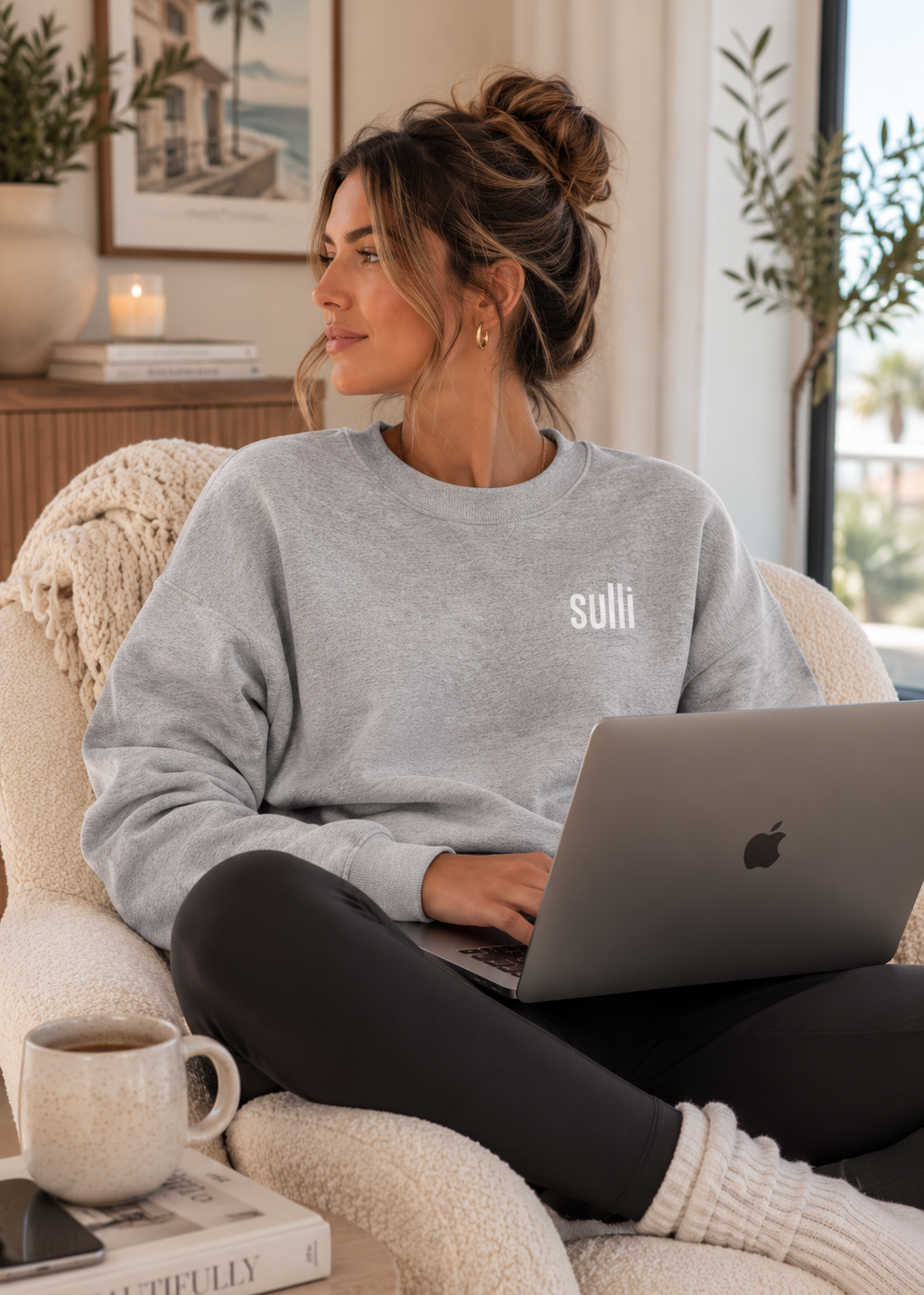 A woman sits in a cozy armchair, wearing a light gray crewneck sweatshirt with the brand name 'sulli' printed in white on the left chest. She is using a silver laptop on her lap in a bright, modern living room setting.