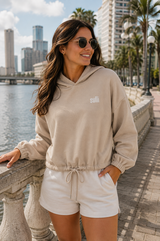 A woman wearing a beige pullover hoodie with a small white 'sulli' logo on the chest, paired with white drawstring shorts, standing on a waterfront promenade with a city skyline and palm trees in the background.