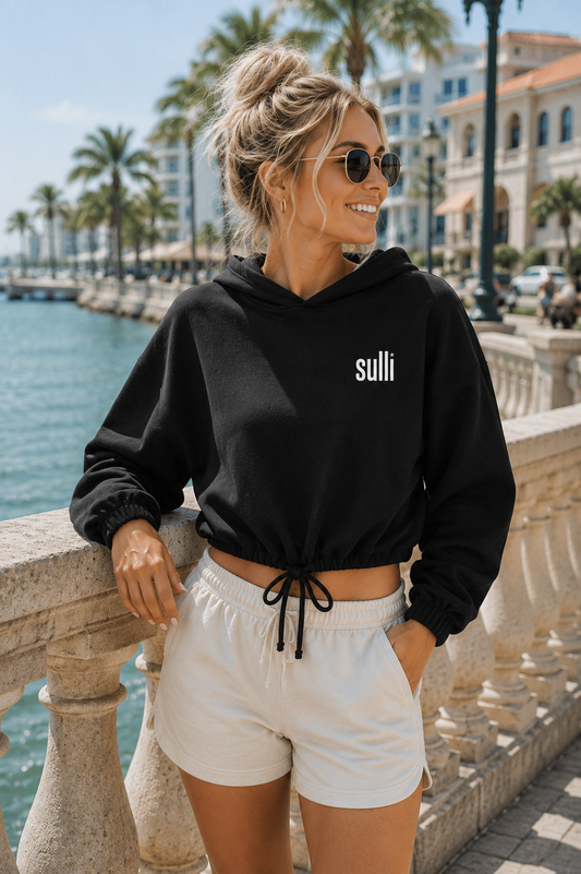A woman wearing a black cropped hoodie with 'sulli' printed in white on the chest, paired with white shorts, posing outdoors by a waterfront with palm trees and buildings in the background.