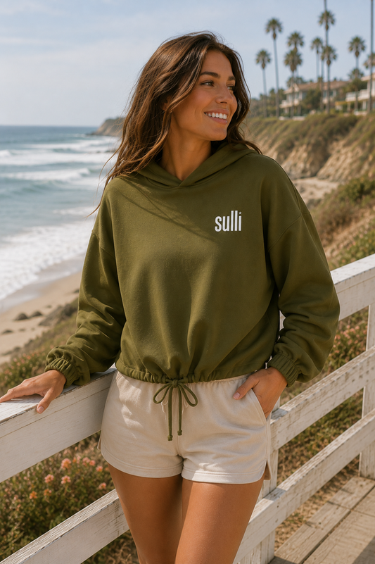 A woman wearing an olive green hoodie with 'suli' printed in white on the chest, paired with cream shorts, posing on a wooden boardwalk overlooking a beach and ocean.