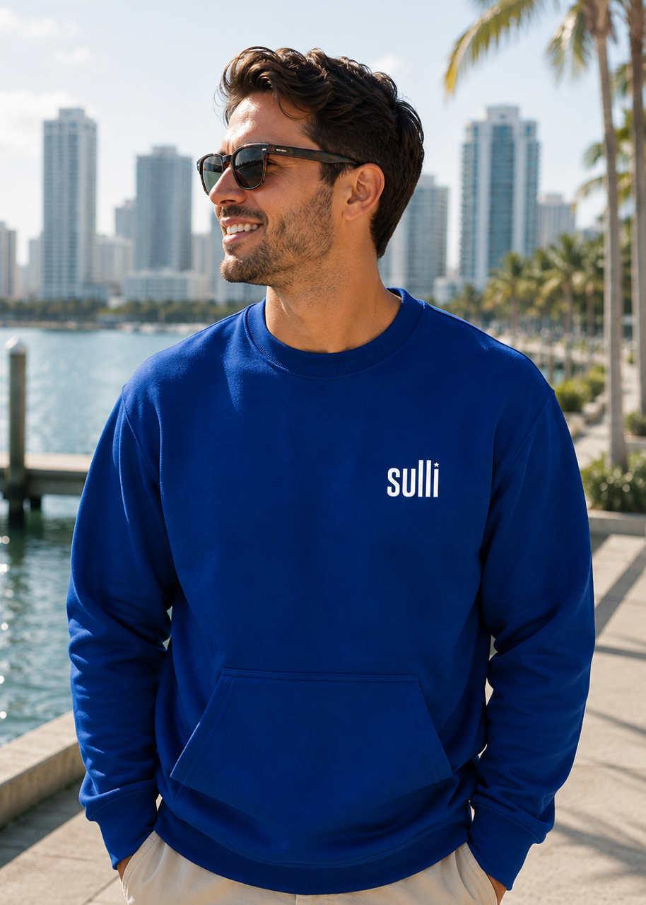 A man wearing a royal blue crewneck sweatshirt with the brand name 'sulli' printed in white on the left chest, paired with beige pants and sunglasses, standing outdoors near a waterfront with a city skyline and palm trees in the background.