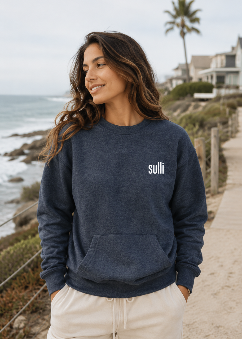 A woman wearing a navy blue crewneck sweatshirt with the word 'sulli' printed in white on the left chest, standing outdoors on a beachside path.