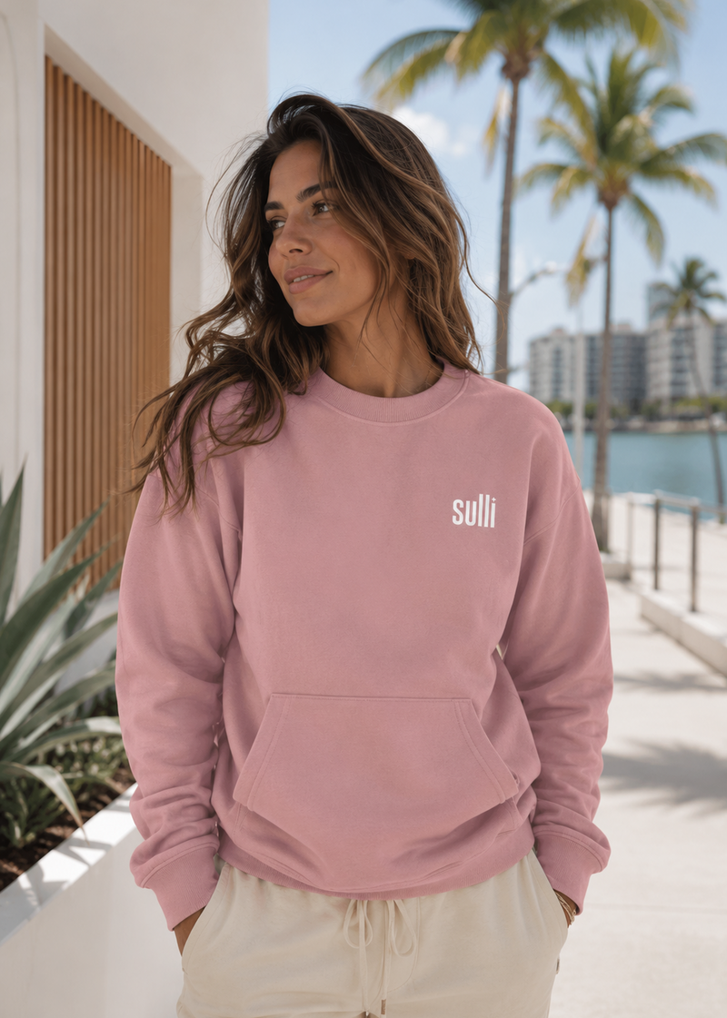 A woman wearing a Sulli Pale Blush crewneck sweatshirt with a white logo, standing along a sunny waterfront with palm trees and a modern city skyline.