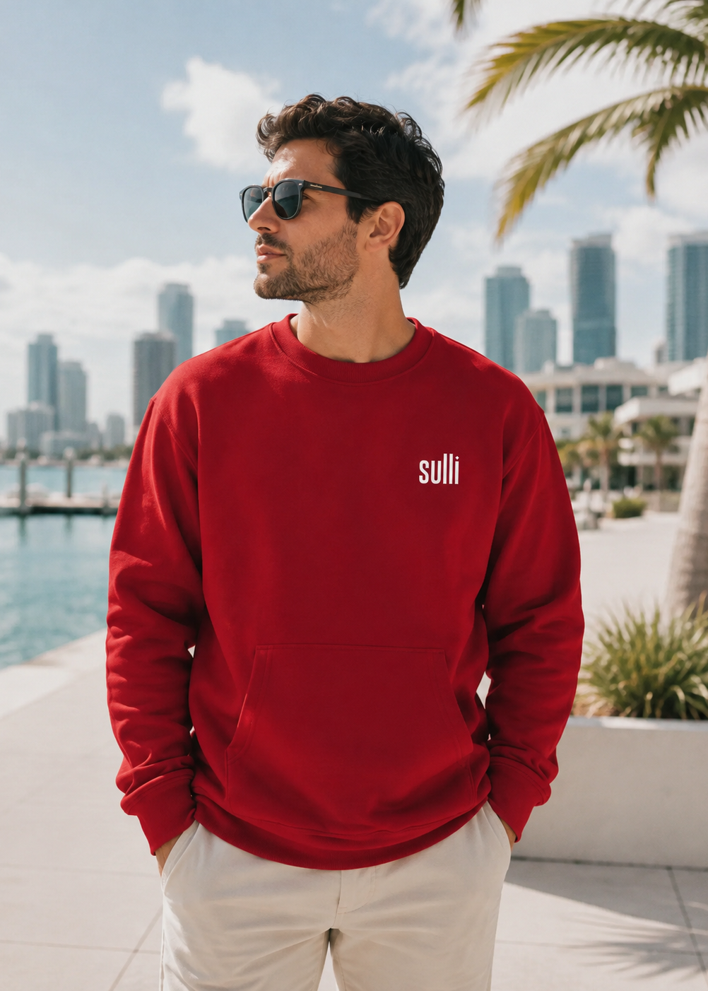 A man wearing a red crewneck sweatshirt with the brand name 'suli' printed in white on the left chest, paired with white pants and sunglasses, standing outdoors near a waterfront with a city skyline and palm tree in the background.