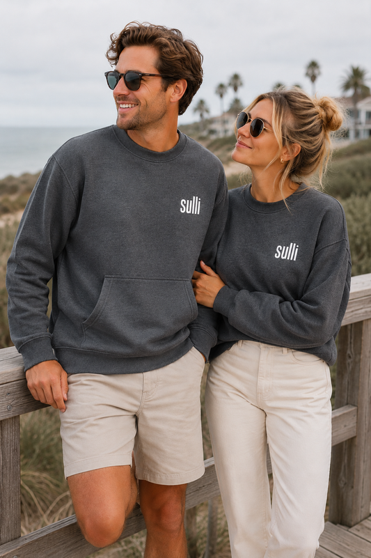 A man and a woman wearing matching dark gray crewneck sweatshirts with the white text 'sulli' on the chest, standing on a wooden boardwalk near the beach.