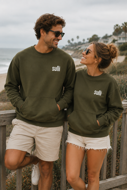 A man and a woman wearing matching olive green crewneck sweatshirts with the white 'sulli' logo on the chest, standing on a wooden boardwalk by the beach.