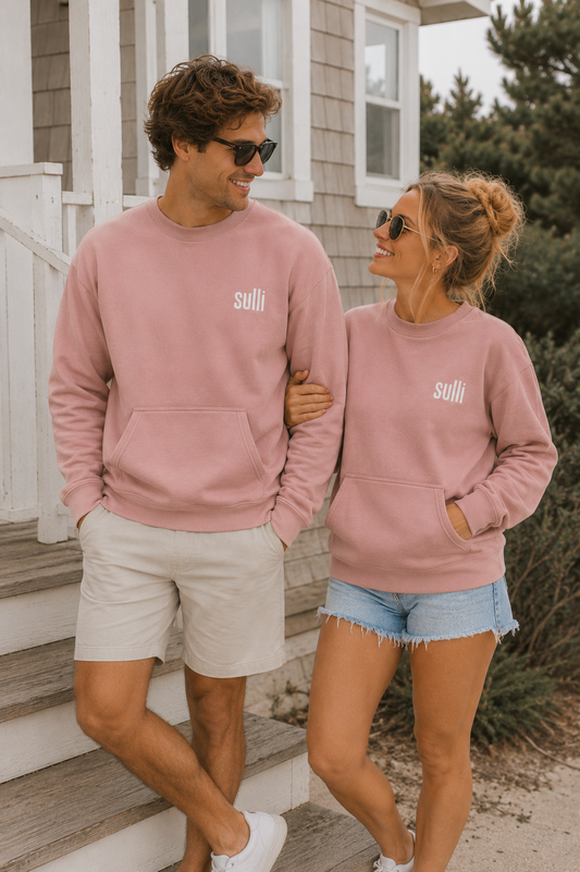 A man and a woman standing on outdoor steps wearing matching dusty rose crewneck sweatpants with the white 'sulli' logo on the left thigh, paired with light shorts and denim shorts.