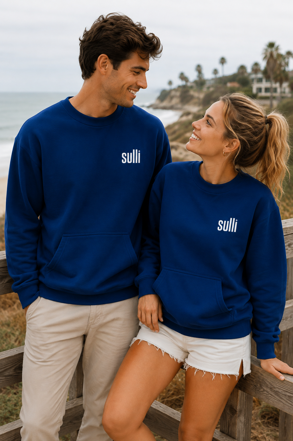 A man and a woman wearing matching navy blue crewneck sweatshirts with the white text 'sulli' on the chest, standing outdoors near a beach and wooden railing.