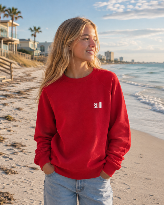 A woman standing on a sandy beach wearing a bright red crewneck sweatshirt with the word 'sulli' printed in white on the left chest, paired with light blue jeans.