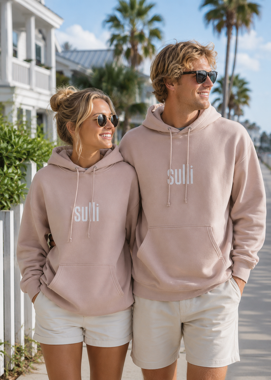 A man and a woman standing together outdoors on a sunny beachside walkway, both wearing matching dusty pink hoodies with the word 'sulli' printed in white across the chest, paired with white shorts.