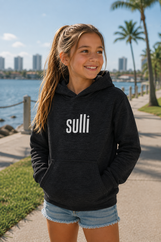A young girl with a ponytail stands on a sunny boardwalk by the water wearing a dark charcoal gray heather hoodie with the word 'sulli' printed in white across the chest, paired with denim shorts.