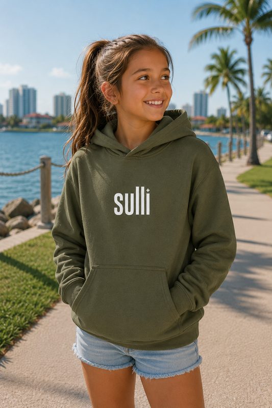 A young girl with a ponytail stands on a waterfront pathway wearing an olive green hoodie with the word 'sulli' printed in white across the chest, paired with denim shorts. Palm trees, water, and a city skyline are visible in the background.