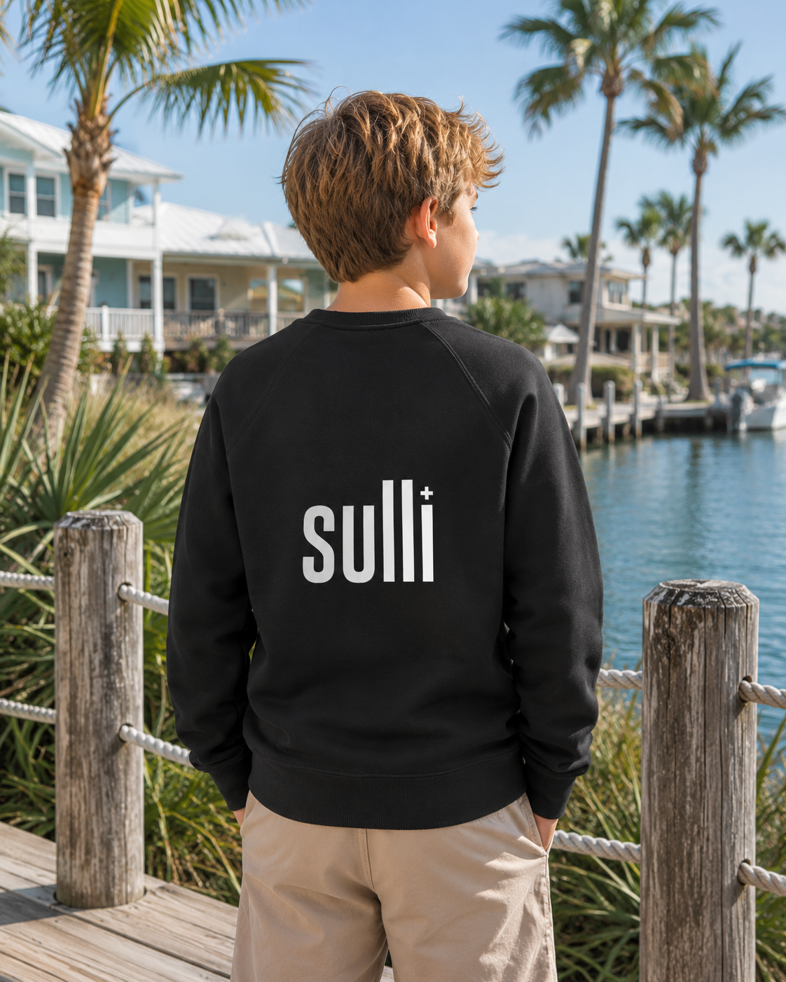 A boy stands on a wooden dock by the water, viewed from behind, wearing a black crewneck sweatshirt with the brand name 'sulli' printed in white across the back, paired with khaki pants.