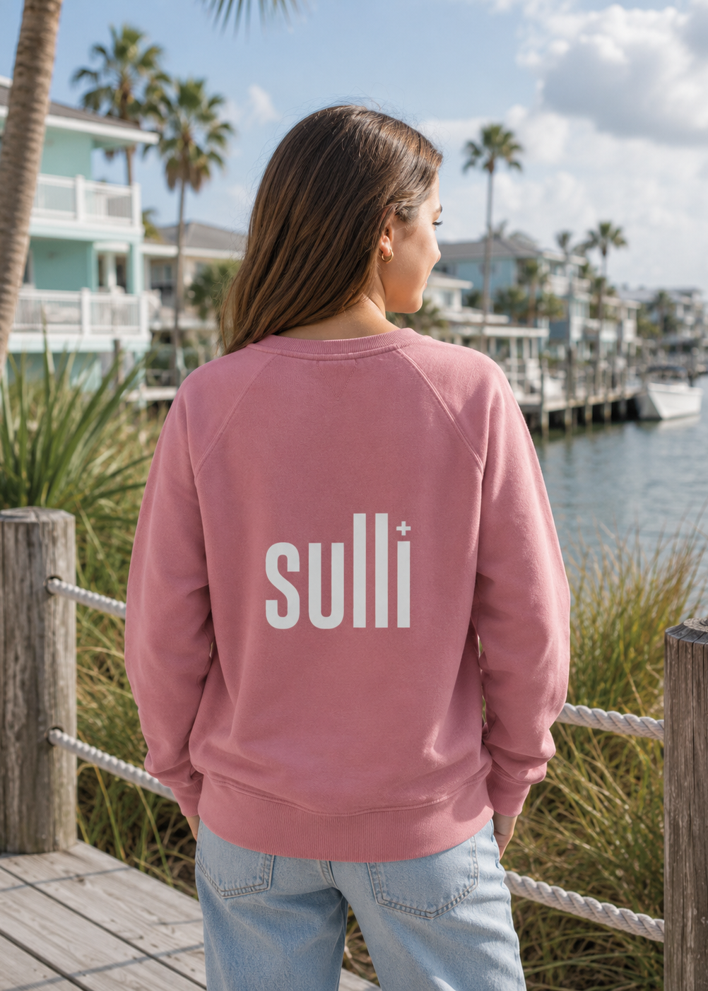 A woman stands on a wooden dock by the water, viewed from behind, wearing a dusty rose crewneck sweatshirt with the brand name 'sulli+' printed in white across the back, paired with light blue jeans.