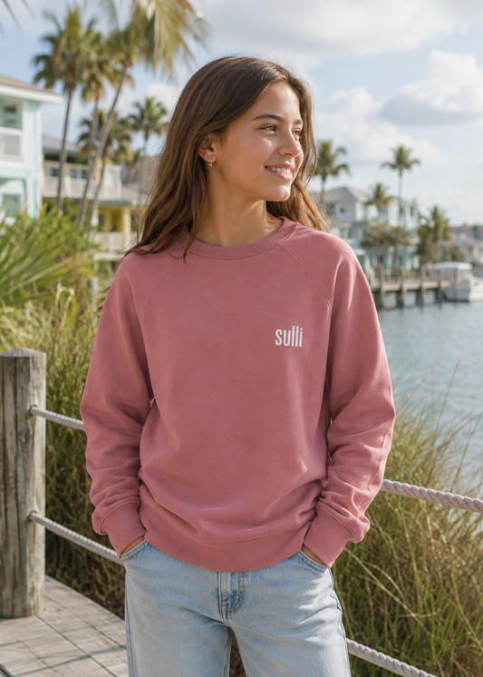A woman wearing a dusty rose crewneck sweatshirt with the brand name 'sulli' printed in white on the left chest, standing outdoors by a waterfront with palm trees and houses in the background.