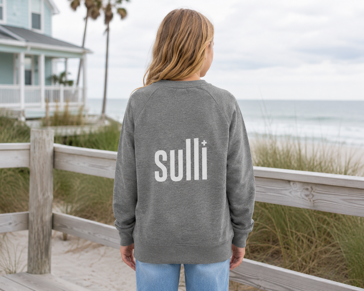 A woman viewed from behind wearing a heather gray crewneck sweatshirt with 'sulli+' printed in white across the back, standing on a wooden boardwalk with a beach and house in the background.