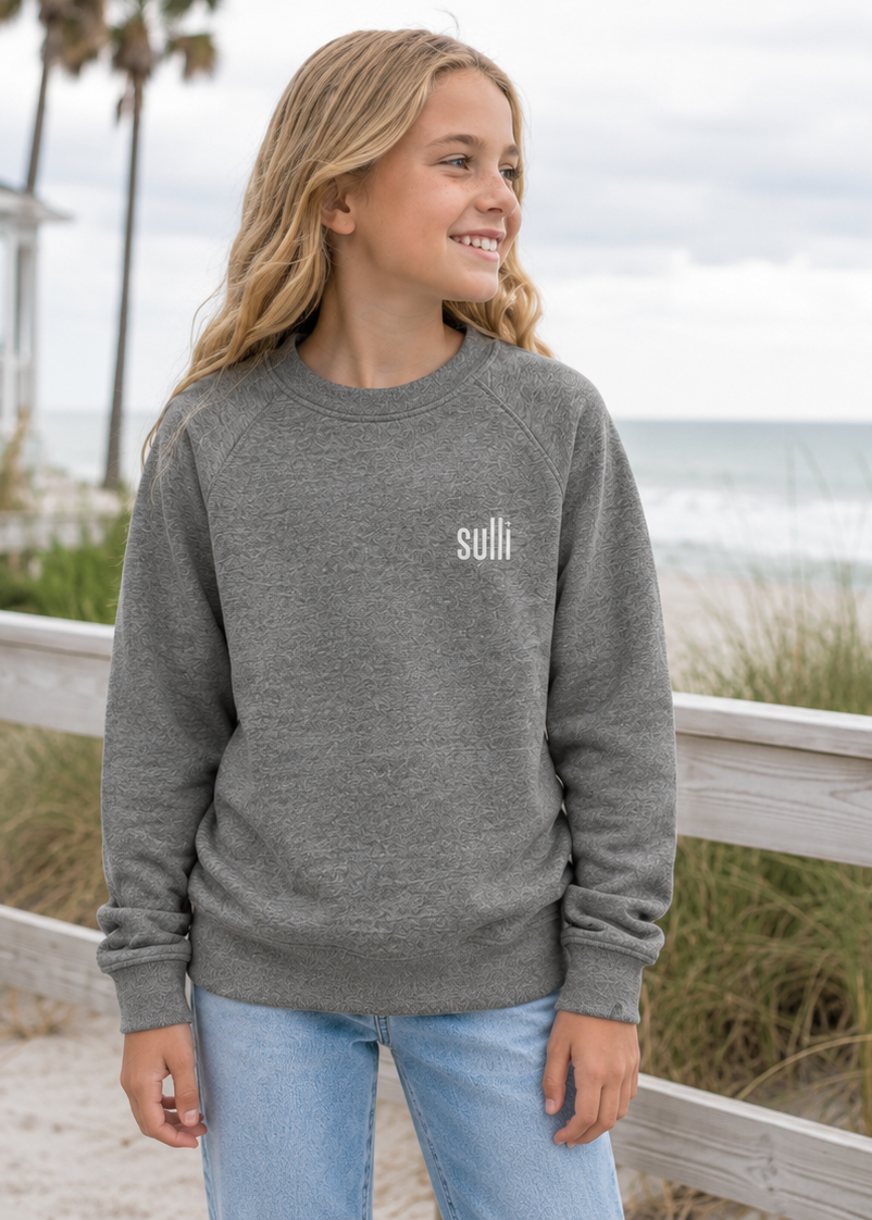 A young woman stands on a beach boardwalk wearing a heather gray crewneck sweatshirt with the brand name 'sulli' printed in white on the left chest, paired with light blue jeans.