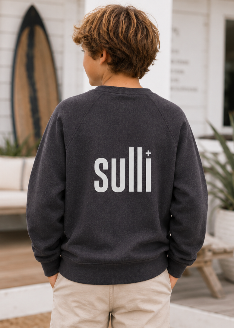 A child viewed from behind wearing a dark charcoal gray crewneck sweatshirt with 'sulli+' printed in white across the back, standing on a beach house porch.