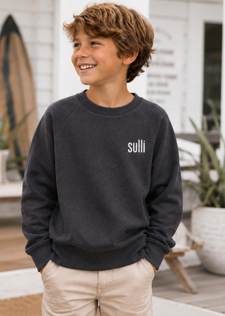 A smiling young boy with curly brown hair stands on a beachside porch wearing a dark charcoal gray crewneck sweatshirt with the brand name 'sulli' printed in white on the left chest, paired with beige pants.